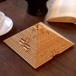 ModÚle de pyramide en bois gravée avec des motifs scientifiques, posé sur une table en bois avec un livre ouvert et une tasse à café à proximité.