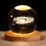 Boule de cristal avec une représentation d'une galaxie en suspension, illuminée et posée sur un socle en bois, idéale pour la décoration scientifique et les amateurs d'astronomie.