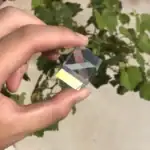 Pierre en verre optique, cristal ou gemme miniature tenue entre les doigts, avec un fond de feuillage vert.