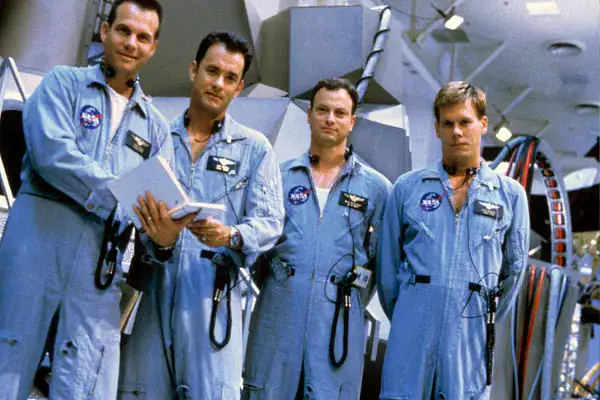 Four astronauts in blue space suits standing inside a spacecraft, preparing for a mission with equipment and control panels in the background.