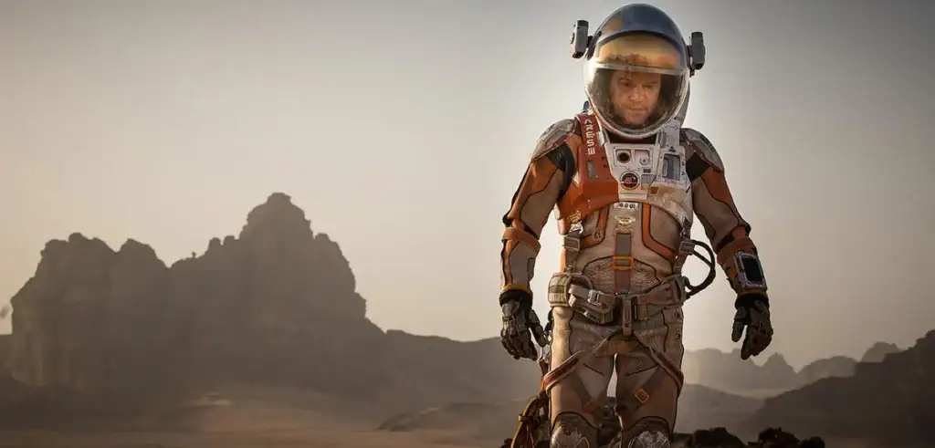 An astronaut in a detailed space suit walking across a rocky, desert-like terrain with mountains in the background under a hazy sky.