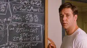A male physics teacher explaining complex equations on a chalkboard filled with mathematical formulas in a classroom setting.