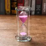 Sablier en verre avec sable rose vif, symbole du temps qui passe, posé sur une surface en bois avec une bibliothèque en arrière-plan.