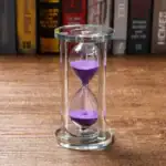 Sablier en verre avec sable violet, symbole du temps qui passe, posé sur une surface en bois avec une bibliothèque en arrière-plan.