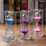 Sablier coloré en verre avec trois tubes contenant du sable bleu, violet et rose, placé sur une surface en bois avec des livres en arrière-plan.