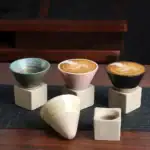 Trois tasses en céramique avec motifs colorés et design moderne, posées sur des supports en béton, sur une surface en bois, illustrant un objet de décoration scientifique.