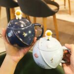 Tasse cosmique avec couvercle et figurine astronaute, décorée de motifs planÚtes, étoiles et lune, idéale pour les amateurs d'astronomie et de décoration scientifique.