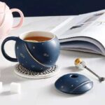 Tasse en céramique bleue avec motifs d'astres, accompagnée d'une souris d'ordinateur assortie, sur un bureau blanc avec un livre ouvert en arriÚre-plan.