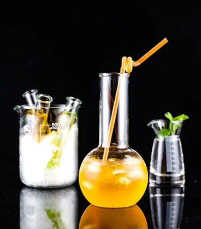 Vase de laboratoire contenant une boisson orange avec une paille, accompagné d'autres accessoires de laboratoire en verre, sur fond noir.