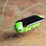 Puce solaire en forme d'insecte avec corps en métal, ailes en plastique noir, et yeux en plastique blanc avec pupilles noires.